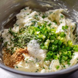 "A close-up shot of a mixing bowl filled with creamy cheese spread, seasoned with fresh herbs like thyme and chopped green onions. The mixture also contains salt, pepper, and spices, giving it a textured, chunky appearance. A spoon rests in the bowl, ready to mix the ingredients further."