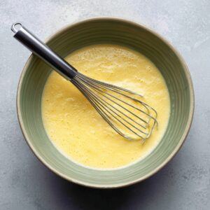 Top-down image of a green ceramic bowl filled with whisked eggs. The eggs are light yellow and frothy, showing fine bubbles on the surface. A metal whisk rests inside the bowl, with egg streaks along the sides, indicating the mixture has just been blended.