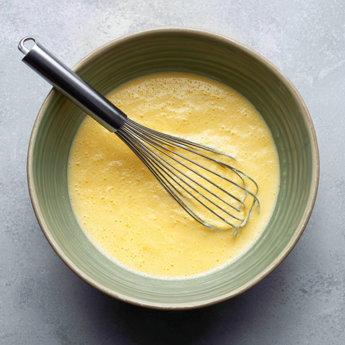 Top-down image of a green ceramic bowl filled with whisked eggs. The eggs are light yellow and frothy, showing fine bubbles on the surface. A metal whisk rests inside the bowl, with egg streaks along the sides, indicating the mixture has just been blended.