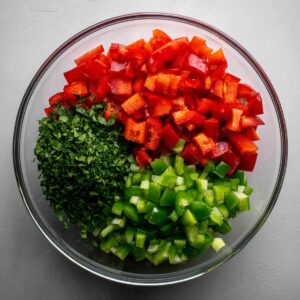 Bright and colorful photo of diced vegetables arranged in sections inside a clear bowl. Includes chopped red bell peppers, green bell peppers, and a generous amount of fresh chopped parsley. The vegetables are vibrant and cleanly chopped, ready to be added to a recipe.