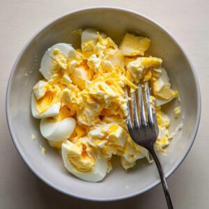 “A fork pressing down on chopped hard-boiled eggs in a bowl as the yolks and whites begin to mash together.
