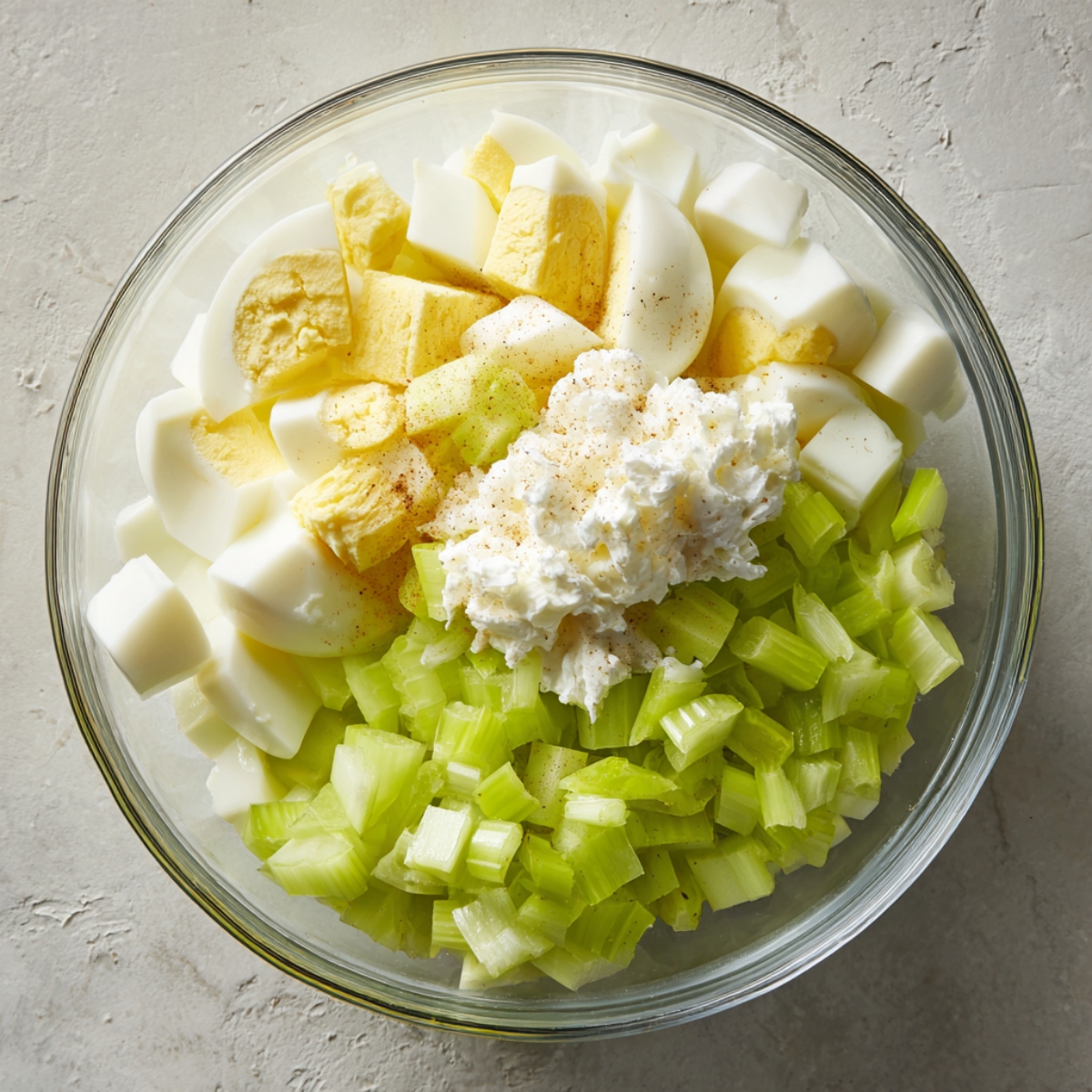 “Mixing bowl holding chopped hard-boiled eggs, diced celery, and a dollop of mayonnaise, set next to a striped tea towel.”