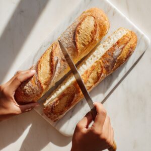 “Overhead shot of a red silicone spatula smoothing a creamy garlic-herb butter across the soft interior of baguette halves, the butter dotted with finely chopped parsley and garlic on a white cutting board.”
