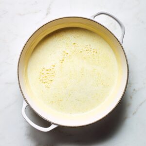 Overhead of a white enameled skillet on marble, filled with uncooked yellow batter, showing tiny bubbles at the edges.