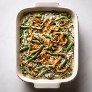 Partially baked green bean casserole in a white ceramic dish, topped with lightly browned crispy onions, fresh green beans, sliced mushrooms, and creamy sauce on a white kitchen counter.
