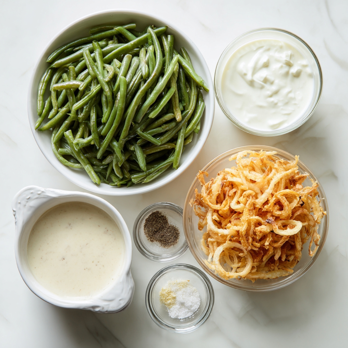 Fresh green beans, creamy soup, sour cream, crispy fried onions, black pepper, salt, and garlic powder arranged in glass and ceramic bowls on a clean marble countertop.
