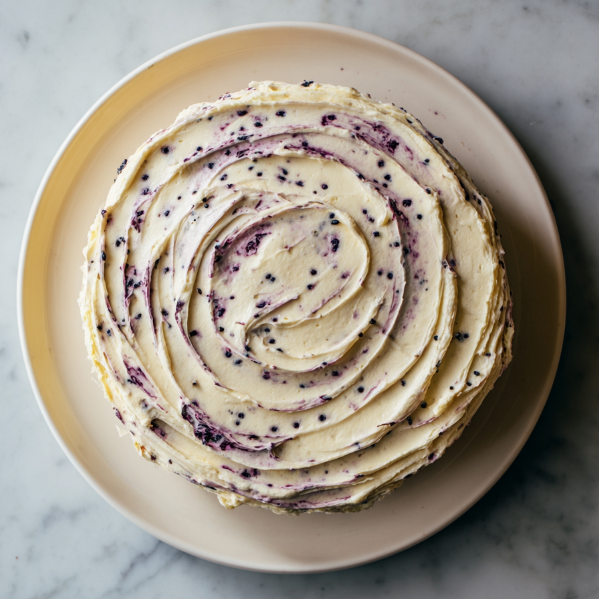 Top-down view of a two-layer lemon blueberry cake completely covered in swirled cream cheese frosting with streaks of deep blueberry-purple swirls. The cake sits on a simple cream-colored plate on a marble countertop, and the frosting’s circular peaks and troughs catch the light.