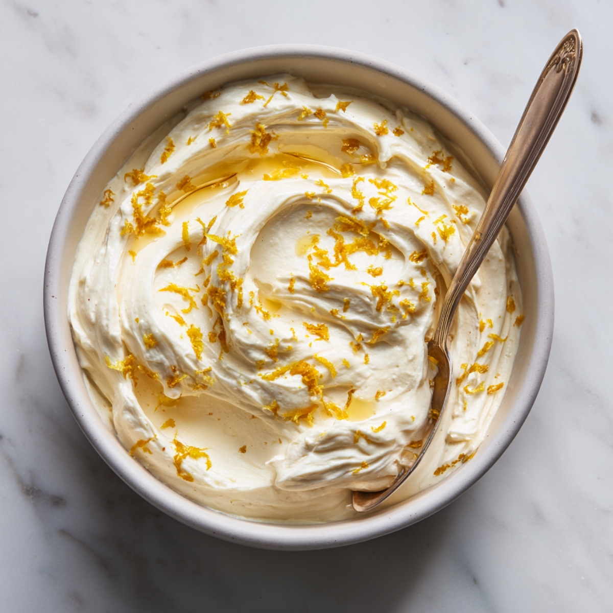 A close-up overhead view of a white bowl filled with creamy lemon frosting, swirled to form peaks and valleys, sprinkled with bright yellow lemon zest, and lightly drizzled with glossy lemon juice. A silver spoon tucks into the side, and soft daylight highlights the frosting’s silky texture.