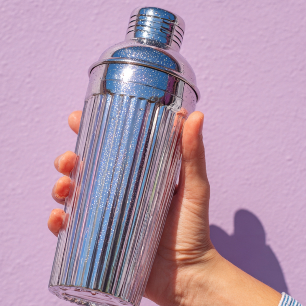 A close-up of a right hand in a striped denim shirt sleeve firmly gripping a tall, fluted-glass cocktail shaker with a metal cap. The shaker’s frosty surface glistens with condensation droplets. A pale lavender wall blurs softly behind. Light from the side window emphasizes the texture of the glass and the hand.
