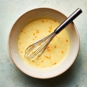 Close overhead view of a speckled ceramic mixing bowl on a light, textured work surface. Inside, a creamy, pale-yellow orange-infused batter flecked with bright zest swirls around a stainless-steel whisk with a matte-black handle. Dappled daylight from the top left creates gentle shadows on the batter’s rippled surface and whisk wires—suggesting someone just paused mid-mix.