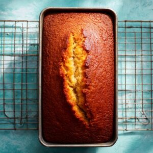 Top-down view of a just-baked orange loaf still in its metal pan, resting on an antique wire cooling rack atop a mottled turquoise tabletop. The cake’s deep golden-brown crust is cracked down the middle, revealing its moist, tender crumb. Soft shafts of daylight from the upper right cast a crisp grid-pattern shadow of the rack and highlight the shiny, slightly syrupy sheen on the crust—evoking a warm, just-out-of-the-oven moment.