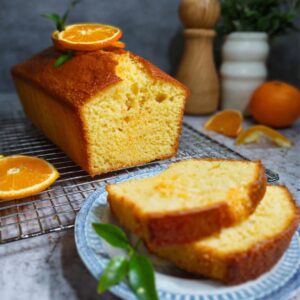 A close-up, side-angle shot of a homemade orange cake recipe sliced into thick pieces on a dark slate board. The front slice reveals a tender, fine-crumb interior and a glossy, amber-hued glaze dripping slightly down its golden-brown crust. Each slice is crowned with a thin, candied orange round sprinkled with bright green zest. In the foreground, a fresh mint sprig rests next to an extra orange wheel, all bathed in soft, natural light that casts gentle shadows and gives the scene a warm, cozy kitchen feel.