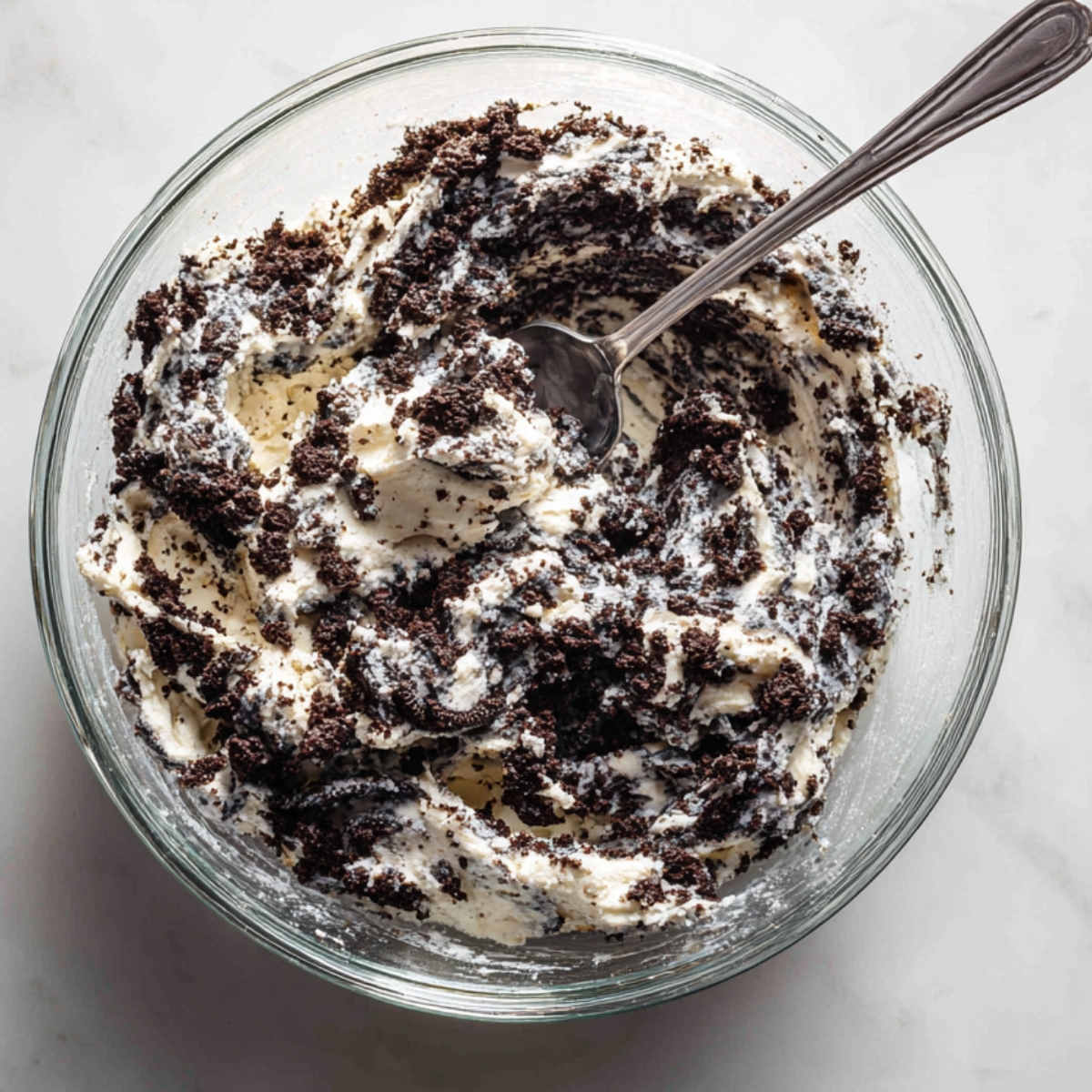 A clear glass mixing bowl with crushed Oreo cookies and softened cream cheese being stirred together. The mixture is half-blended, with visible dark crumbs and white swirls, and a spoon resting in the bowl, all on a white marble surface.