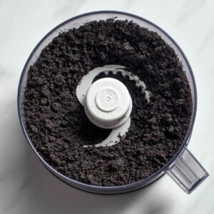 A top-down view of a food processor bowl filled with finely ground Oreo cookie crumbs. The texture is uniform and dark, sitting around a white plastic blade insert on a clean marble countertop.