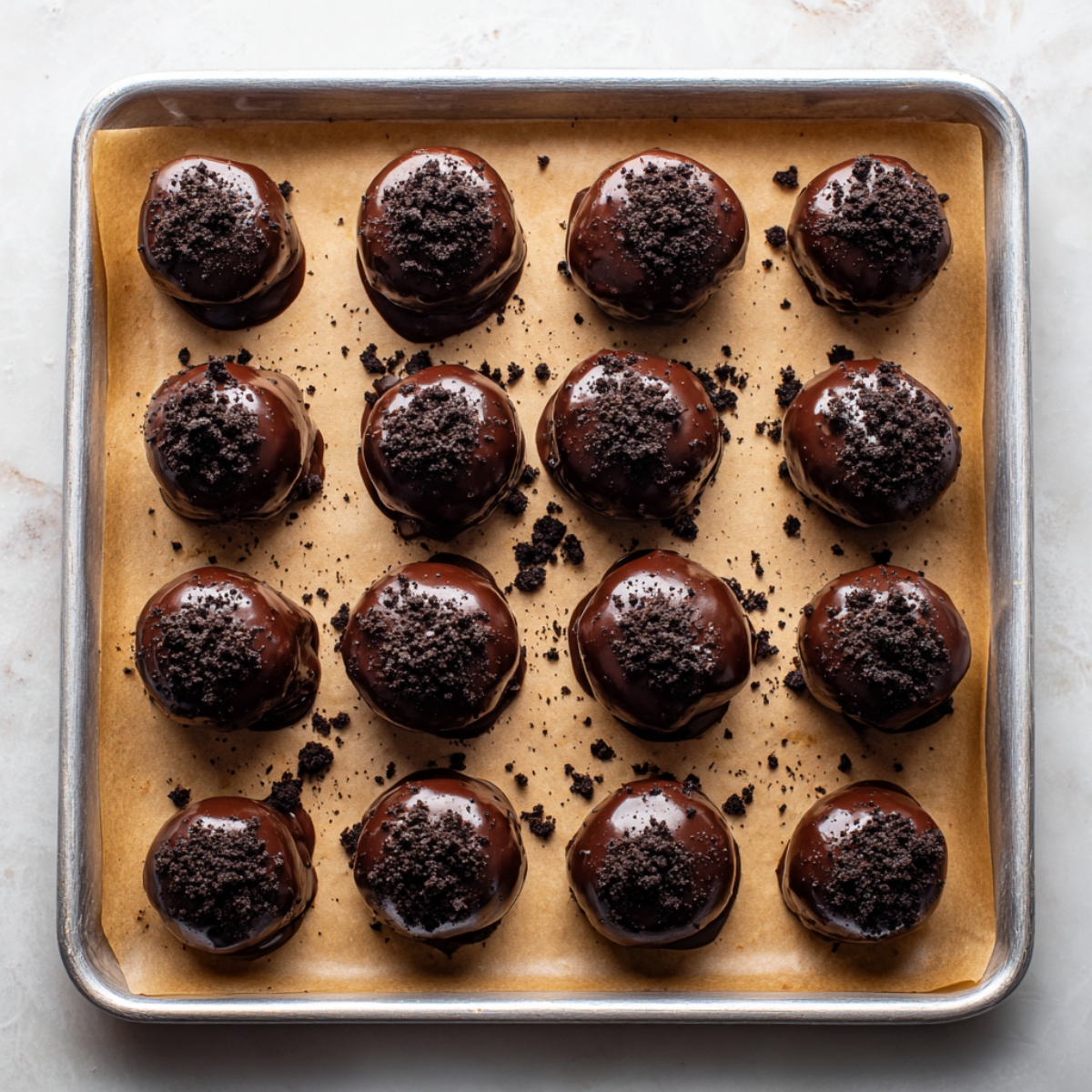 A clean, parchment-lined baking sheet with perfectly arranged chocolate Oreo truffles in a 4x4 grid. Each truffle is smooth and glossy, generously topped with crushed cookies, with crumbs scattered around for a natural, homemade vibe.