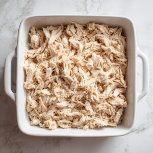 “Overhead view of a square white ceramic casserole pan with handles, completely filled with an even layer of freshly shredded chicken breast. The pale, fibrous strands cover the bottom of the dish, resting on a marble countertop, bathed in clear natural light that accentuates each individual shred.”