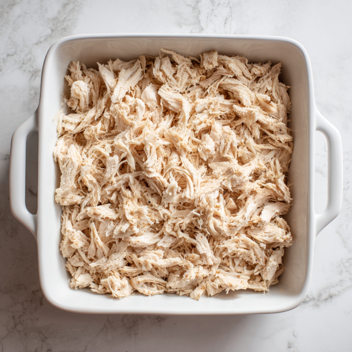 “Overhead view of a square white ceramic casserole pan with handles, completely filled with an even layer of freshly shredded chicken breast. The pale, fibrous strands cover the bottom of the dish, resting on a marble countertop, bathed in clear natural light that accentuates each individual shred.”
