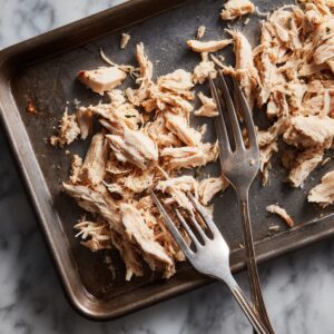 “A birds-eye view of two vintage silver forks pulling apart moist, white shredded chicken on a dark metal baking tray. The flaky strands of chicken are scattered across the tray’s surface, with a few browned bits clinging to the meat. The tray sits on a white marble countertop, and soft, diffused daylight highlights the texture and moisture of the chicken.”