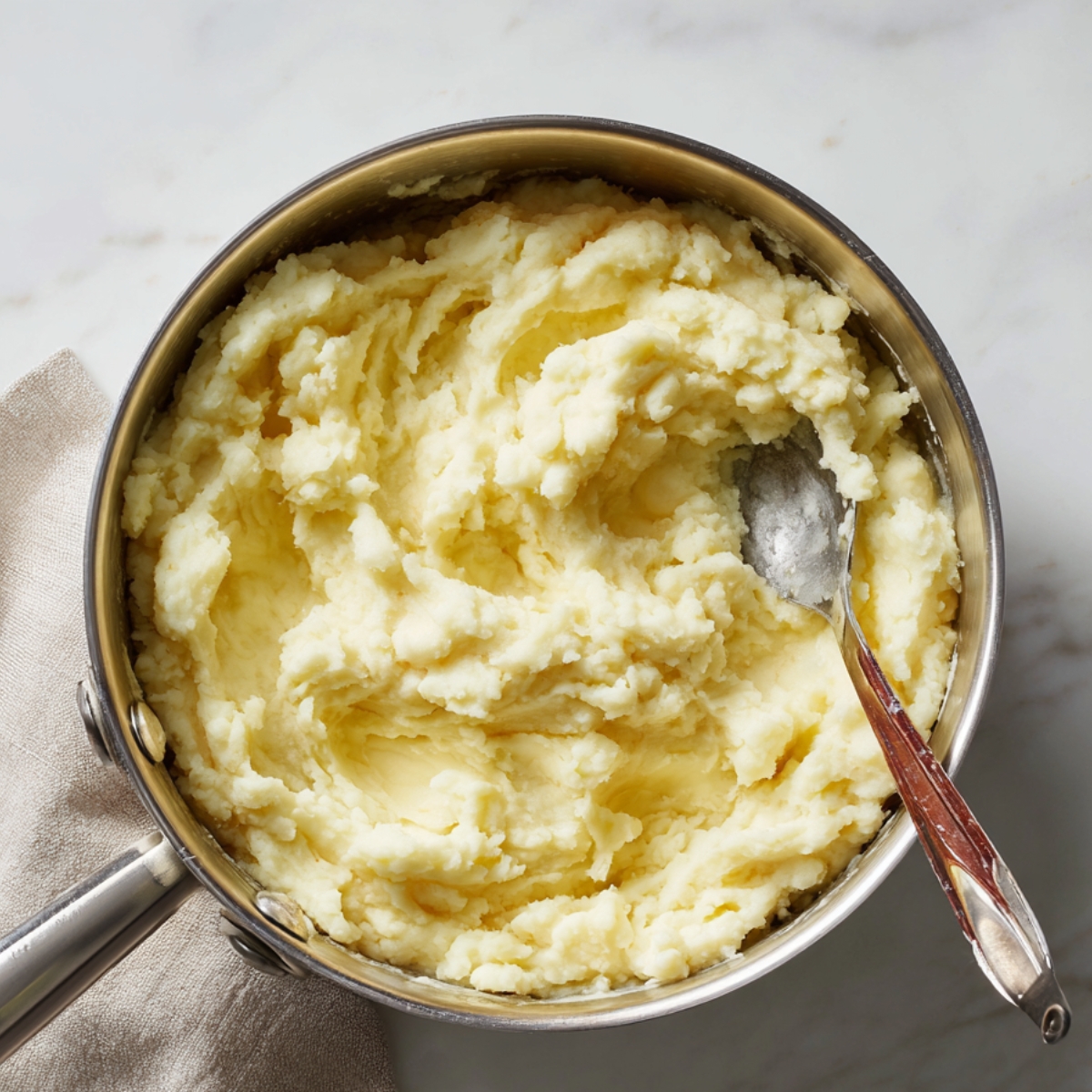 Homemade mashed potatoes in a stainless steel pot with a spoon, resting on a beige cloth over a marble counter. The potatoes look creamy and buttery with a rustic, hand-mashed texture.