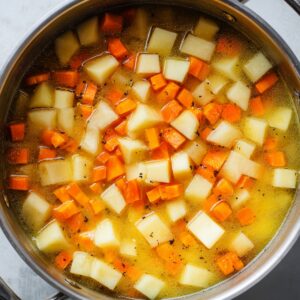 Close overhead of a pot where pale cream is being stirred into tender potato and carrot pieces, creating a smooth, ivory‑colored soup base with small vegetable flecks.
