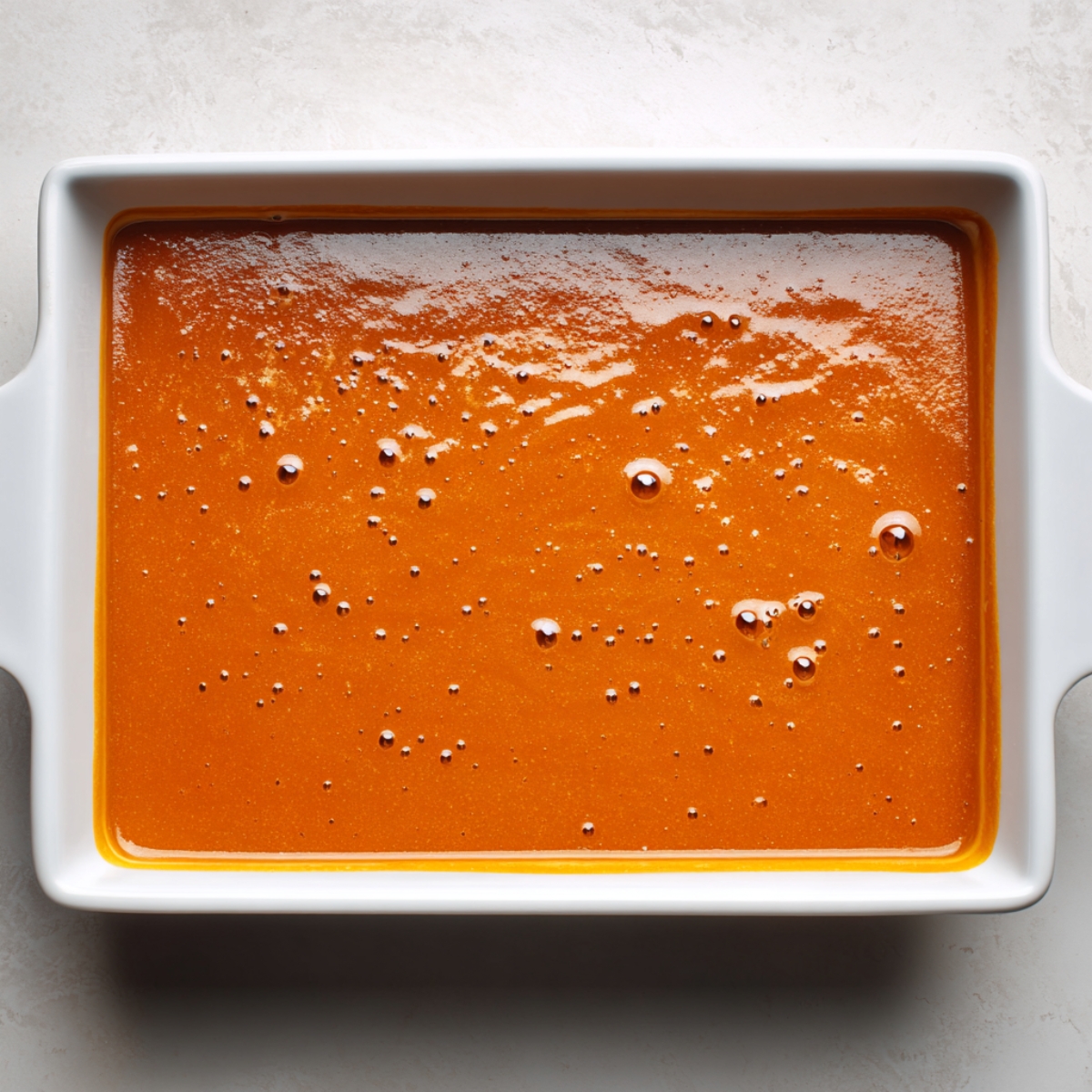A rectangular white baking dish filled with smooth pumpkin pie filling. The surface is glossy, orange, and even, with small air bubbles dotting the top. The dish is placed on a white marble counter, ready for the next layer.