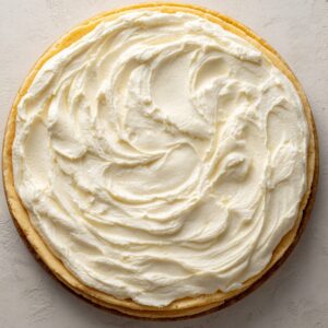 A thick layer of whipped cream cheese frosting swirled smoothly on top of a round cheesecake, presented on a light textured surface in natural light.