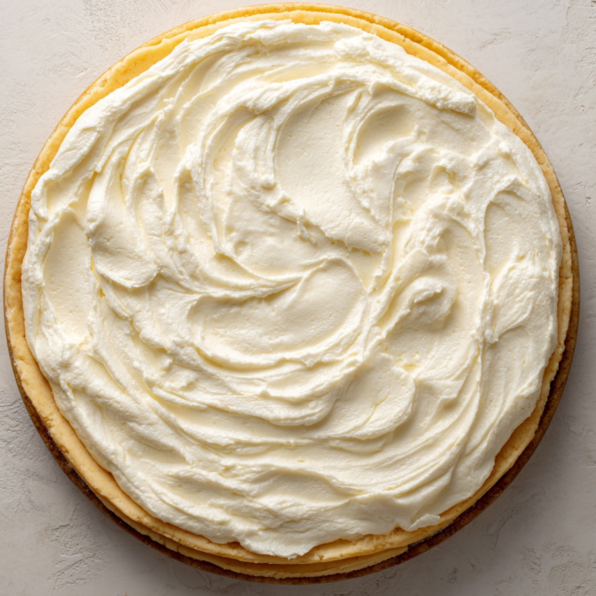 A thick layer of whipped cream cheese frosting swirled smoothly on top of a round cheesecake, presented on a light textured surface in natural light.