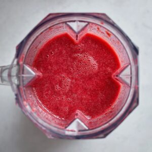 Freshly blended strawberries in a clear blender, smooth red purée with bubbles on top, seen from above.