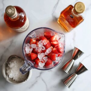 An overhead shot of a blender pitcher now filled with a smooth, bright-red strawberry slush, its surface swirled into a gentle spiral, all sitting on a white marble countertop with soft shadows.