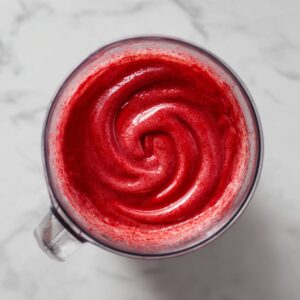 A bird’s-eye view of a clean white marble countertop displaying cocktail prep: a small white bowl of pale-green fresh lime juice, a shallow dish ringed with coarse sea salt, three halved and juiced limes, and the black base of a Vitamix blender, laid out neatly under soft daylight.