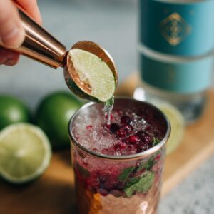 An overhead close-up of a copper jigger tipping freshly squeezed lime juice into the muddled strawberry-mint glass, with whole limes and a bottle of rum blurred behind.