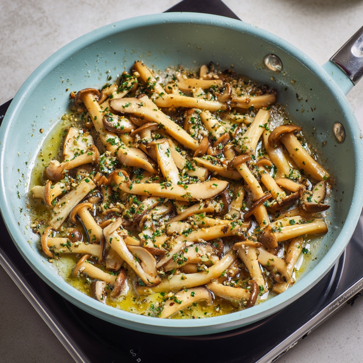 Close-up of a light blue nonstick sauté pan on a black induction cooktop. Minced mushroom stems and diced garlic are bubbling in melted butter, with flecks of chopped herbs visible.