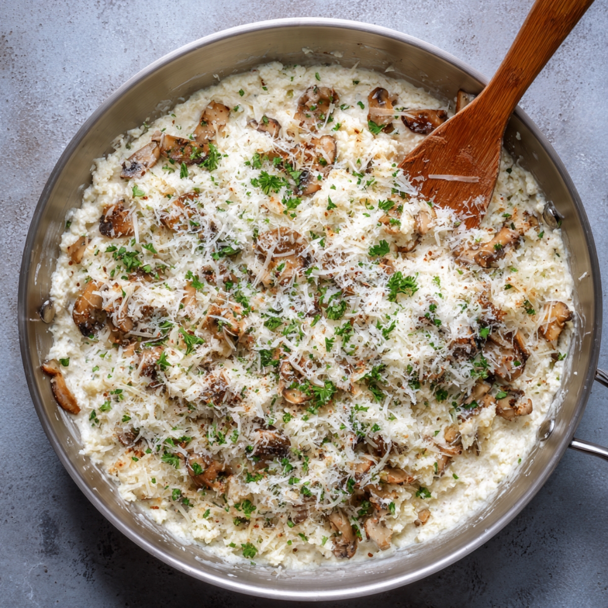 Overhead shot of the same light blue sauté pan, now filled with a thick, ivory-white mixture of cream cheese, grated Parmesan, sautéed mushroom bits, garlic, and chopped parsley being stirred with a wooden spatula.
