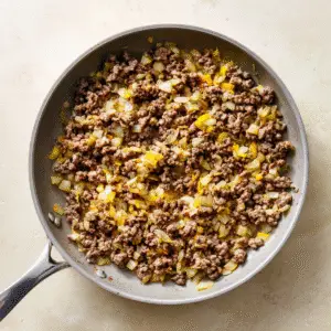 Overhead view of a light-gray nonstick skillet containing freshly browned, coarsely crumbled ground beef mixed with translucent, golden-edged diced yellow onion. The skillet’s stainless-steel handle points toward bottom left, and tiny flecks of cooking oil and browned bits cling to the pan’s interior. It rests on a warm beige stone countertop with subtle mottling, and soft natural light from the top right casts gentle shadows, evoking a just-cooked, homemade moment.