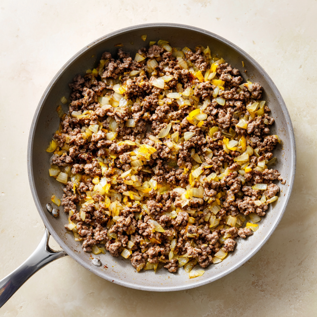 Overhead view of a light-gray nonstick skillet containing freshly browned, coarsely crumbled ground beef mixed with translucent, golden-edged diced yellow onion. The skillet’s stainless-steel handle points toward bottom left, and tiny flecks of cooking oil and browned bits cling to the pan’s interior. It rests on a warm beige stone countertop with subtle mottling, and soft natural light from the top right casts gentle shadows, evoking a just-cooked, homemade moment.