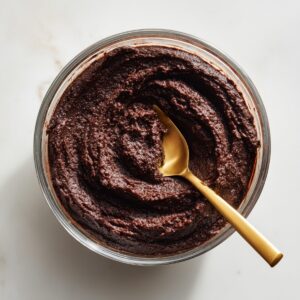 A square baking pan lined with crumpled parchment paper, filled with smooth and evenly spread chocolate brownie batter. The surface has a slight swirl texture, and the pan is placed on a soft-textured, off-white countertop. Looks freshly prepped and oven-ready.
