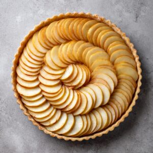 Flat-lay of a fully assembled, unbaked tarte aux pommes: concentric rings of thin apple slices neatly arranged in the raw pastry shell on a light gray countertop.