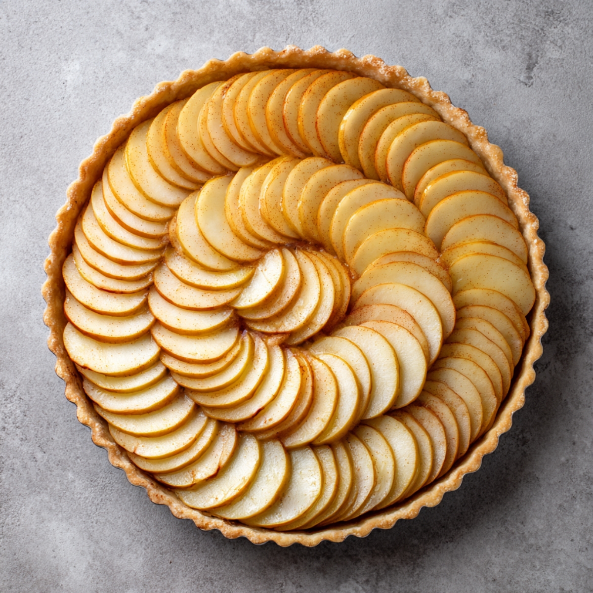 Flat-lay of a fully assembled, unbaked tarte aux pommes: concentric rings of thin apple slices neatly arranged in the raw pastry shell on a light gray countertop.