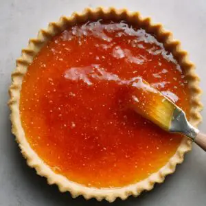 Top-down shot of a fluted pastry shell being brushed with glossy apricot jam by a pastry brush, the vibrant orange glaze evenly coating the bottom.