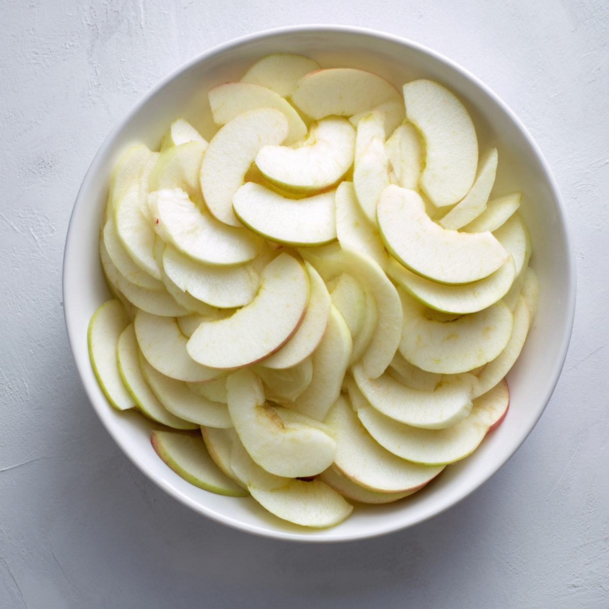 Overhead view of a white bowl piled with thin, peeled apple wedges ready for arranging in the tart, set on a textured white surface.