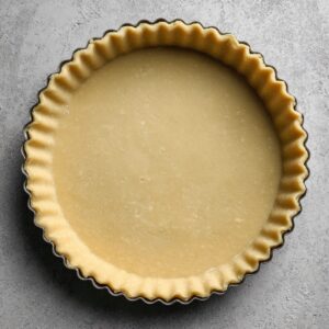 Overhead of an unfilled pâte brisée pressed into a fluted tart pan, the smooth, pale pastry lining ready for the apple filling, on a mottled gray work surface.