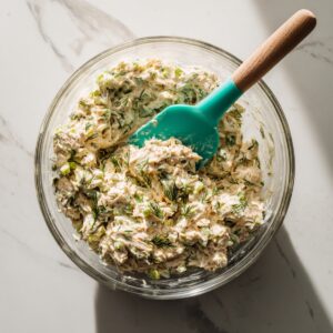 A clear glass mixing bowl on a white marble countertop, containing creamy tuna salad flecked with green celery and herbs, with a teal-handled silicone spatula resting in the bowl under soft natural light.