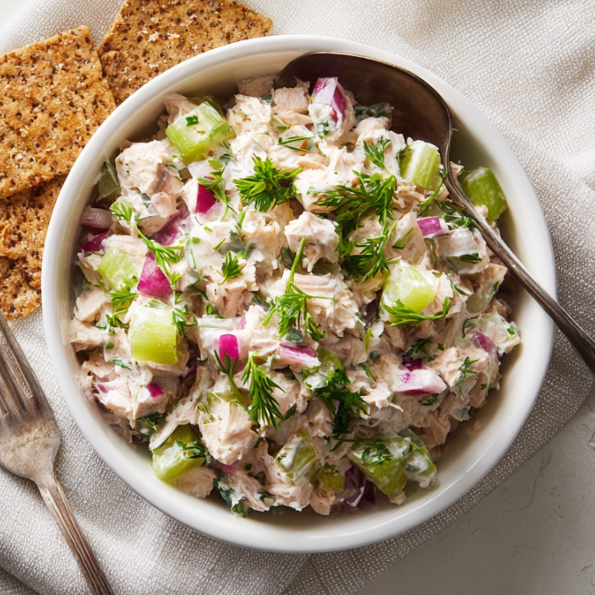 A white ceramic bowl filled with chunky tuna salad—mixed with diced celery, red onion, and fresh dill—served alongside whole-grain crackers on a textured beige napkin, with a silver serving spoon peeking into the frame.