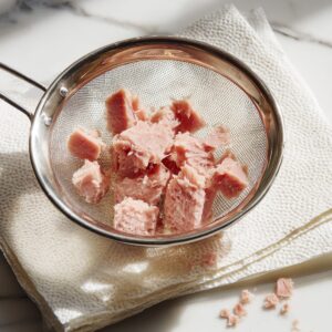 A stainless-steel mesh strainer holding pink tuna chunks over folded paper towels on a white marble countertop, with a few stray flakes of tuna scattered around the base.