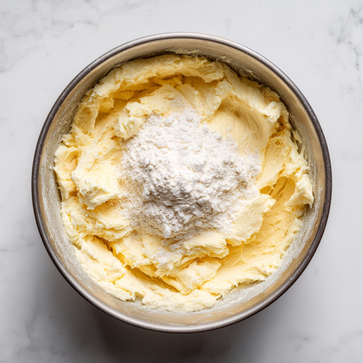 An overhead view of a stainless-steel mixing bowl on a white marble counter, showing pale yellow creamed butter and sugar with a mound of sifted flour and a pinch of baking powder on top, ready to be folded in.