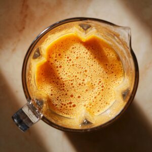 Bird’s-eye view into a clear blender pitcher half-full of smooth, frothy orange carrot purée, the creamy liquid clinging to the sides under warm kitchen light.
