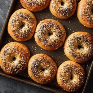 Golden brown homemade bagels fresh out of the oven, topped with a generous blend of sesame, poppy seeds, garlic, and onion. They sit on a dark metal baking tray with baked-on spots, giving a rustic, real-kitchen look.