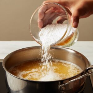 Pouring a white cornstarch slurry from a glass measuring cup into gently bubbling broth in a stainless-steel saucepan.