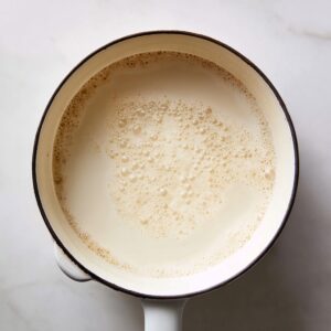 “Top-down shot of a white enamel saucepan with a wooden handle on a white marble countertop, containing smooth whole milk warming to a gentle simmer. Tiny bubbles cluster at the edge, and natural light softly illuminates the creamy surface.”