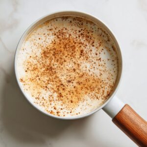 “Overhead view of the same white enamel saucepan on marble, now filled with creamy milk foam sprinkled with ground cinnamon. The foam forms a mottled pattern of white and warm brown, with the wooden handle pointing toward the bottom edge.